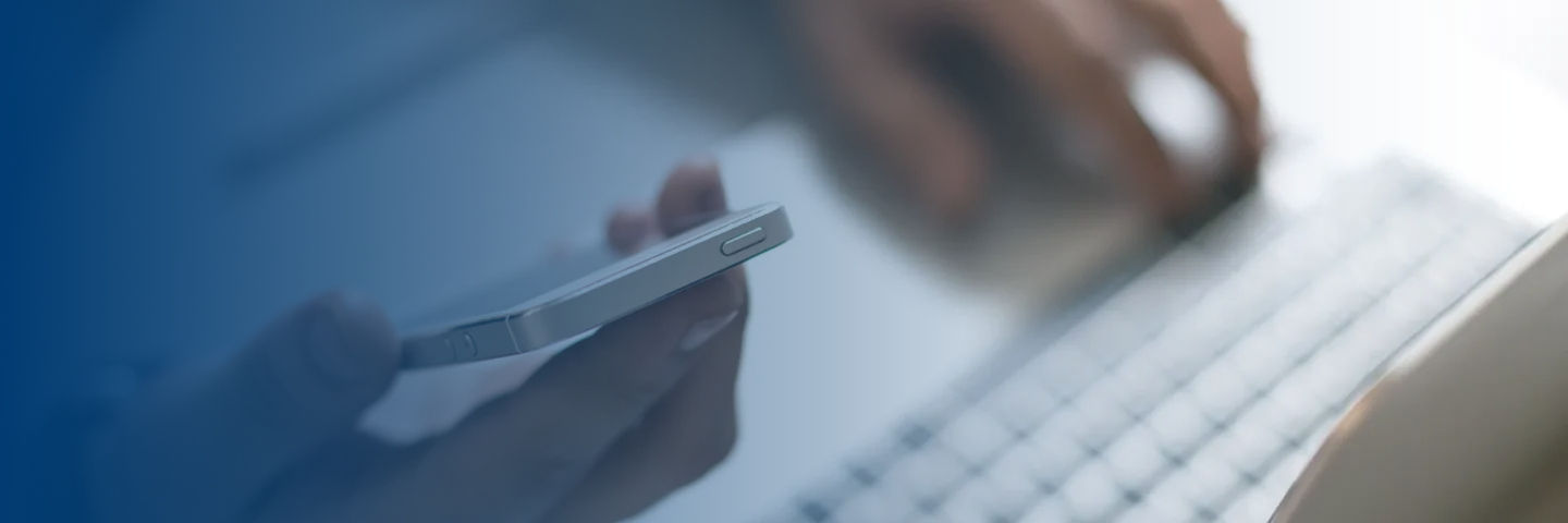 Close-up of a person holding a smartphone with one hand and using a keyboard with the other.